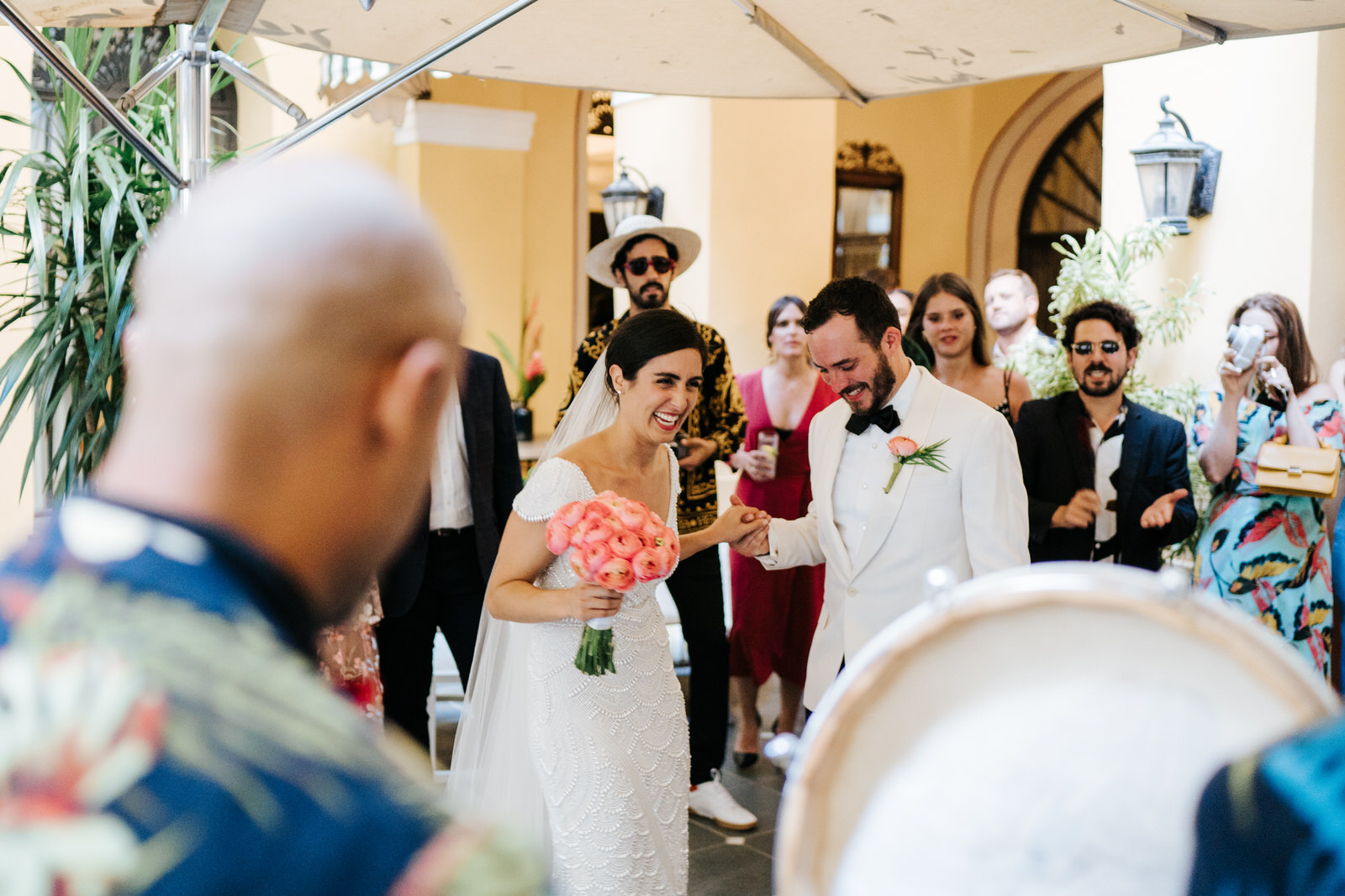 Musicians are playing in the hotel's courtyard as bride and groom hold hands and smile, surrounded by guests