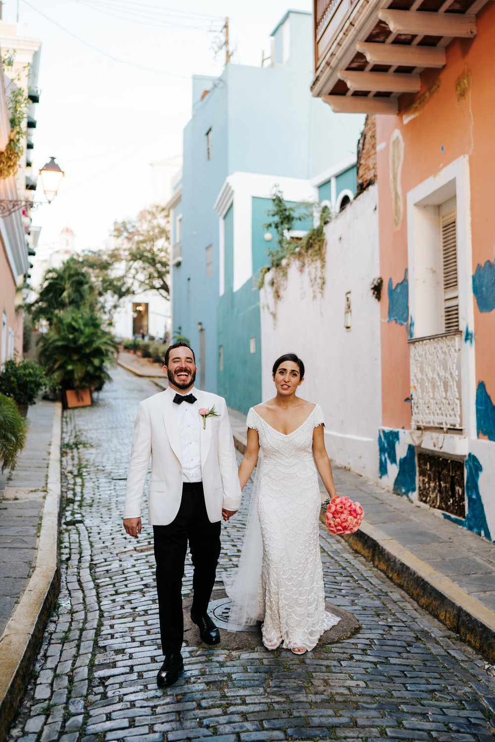  Bride and groom walk down the streets of Old San Juan while groom cannot contain his laughter and bride looks funnily at photographr 