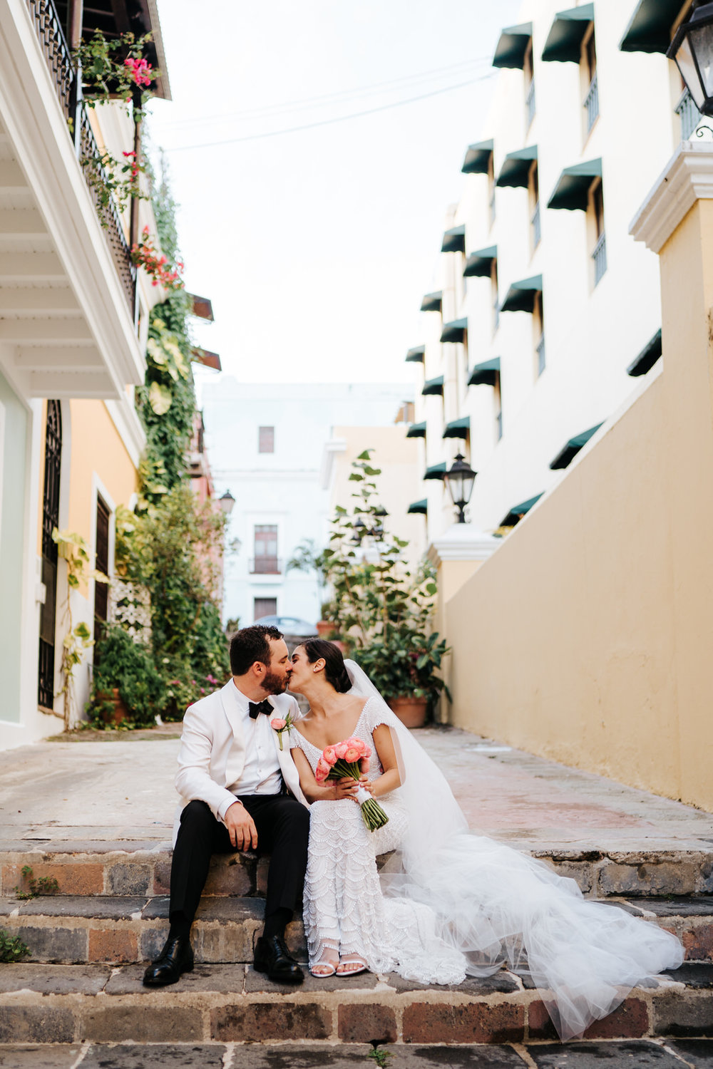  Bride and groom sit on cobblestone steps in an alley in Old San Juan while they kiss 