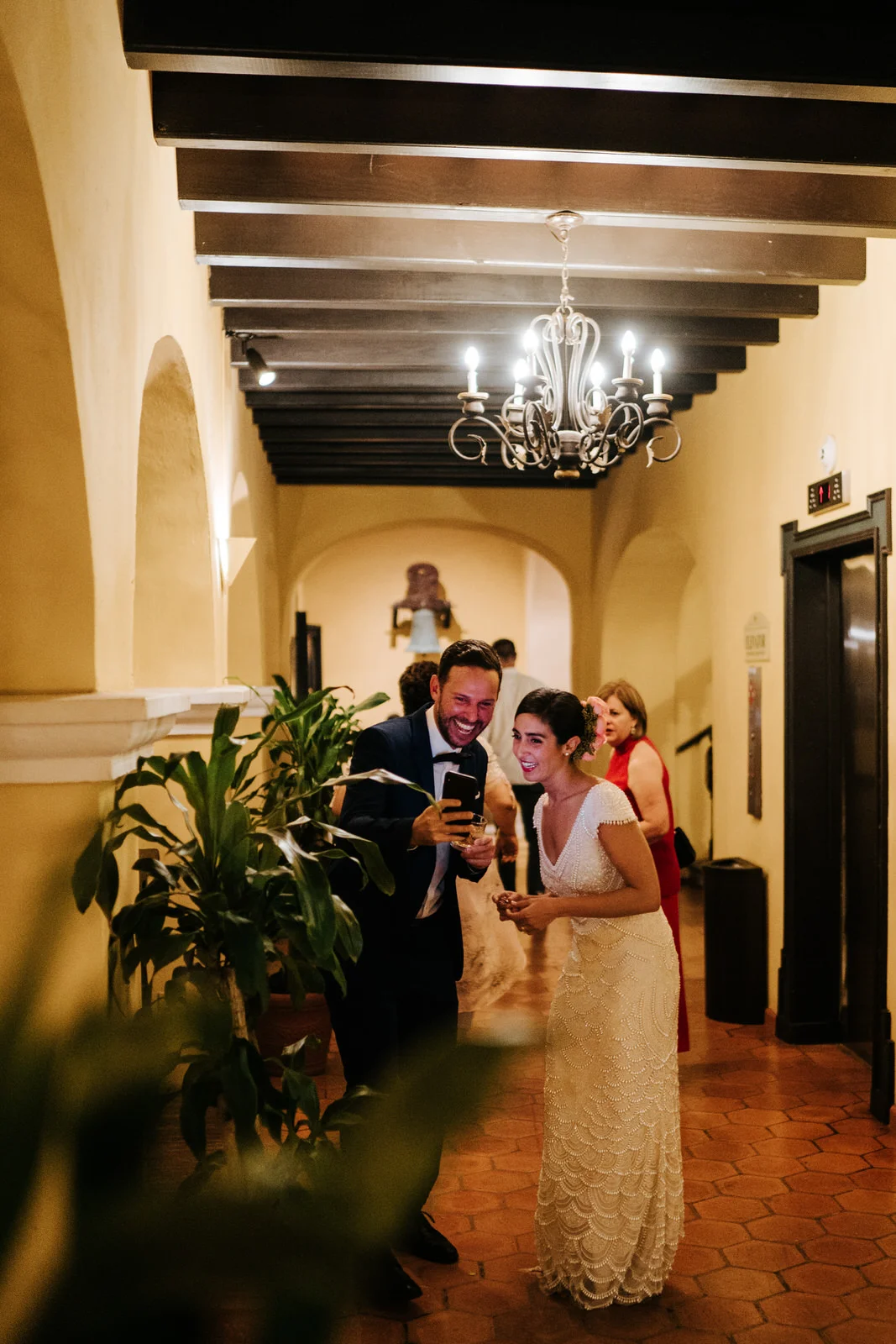 Bride stands in the hallway of the wedding venue as a friend shows her a photo on his phone and they both smile