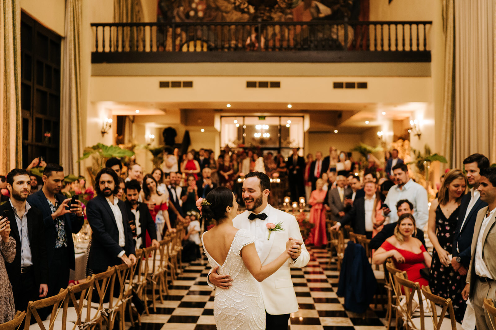 Wide photograph of bride and groom having their first dance. Bride's back is towards the camera while groom smiles at her.