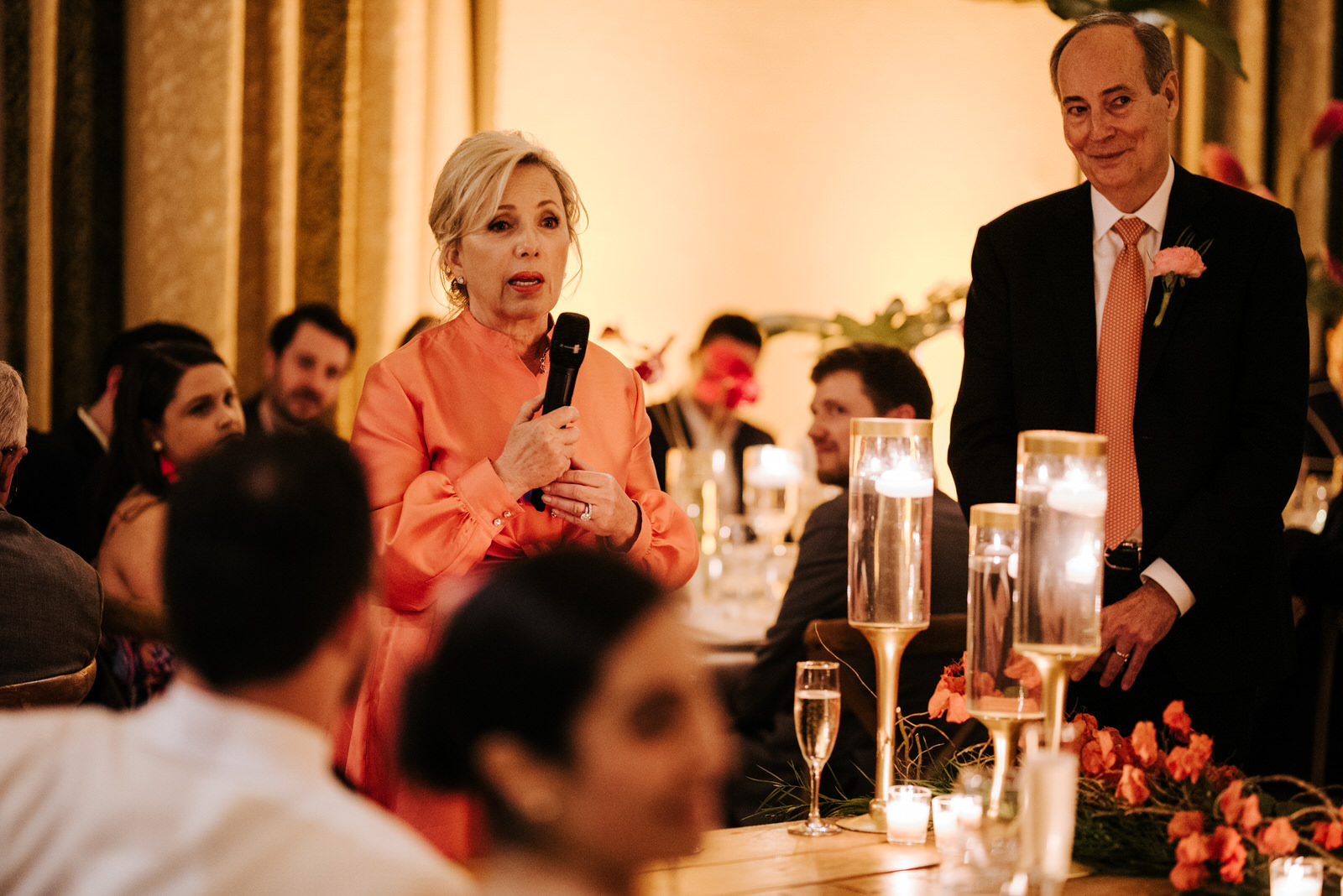 Bride and groom sit in the foreground, out of focus, as the groom's parents do their wedding speech