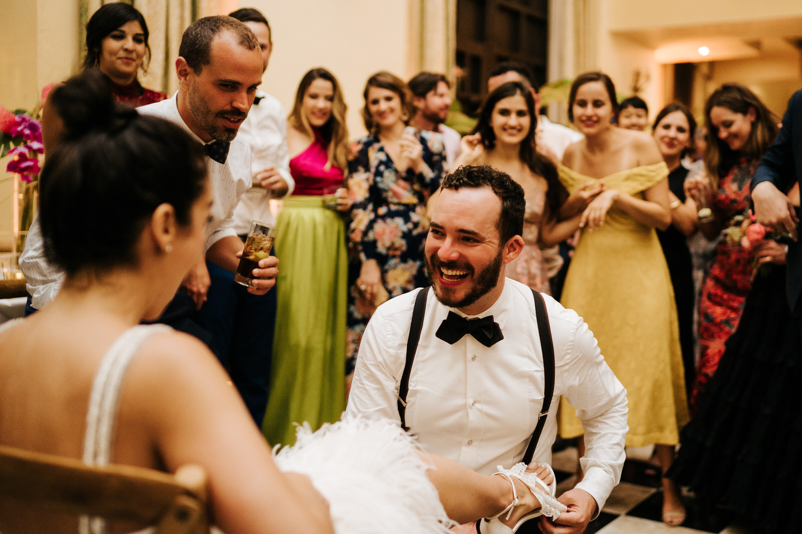 Groom smiles as he takes off the bride's garter as she sits on a chair