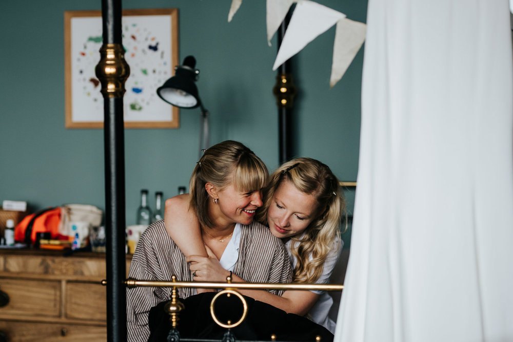  Bride's sister and best friend sit on bed, hugging and smiling at each other 