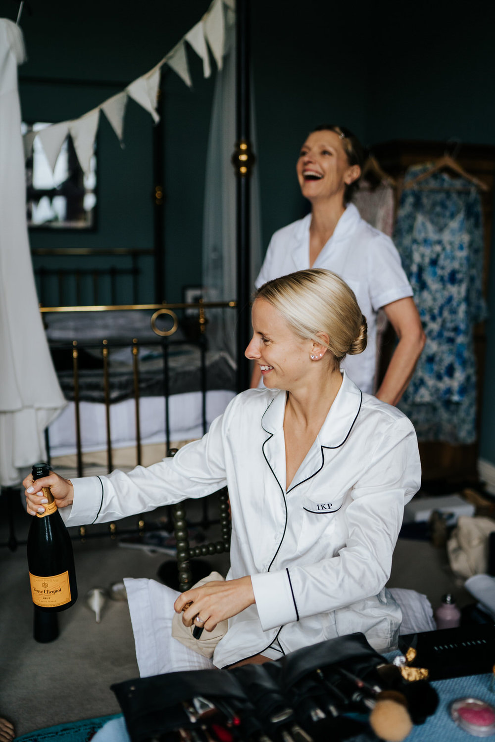  Bride holds and opens a bottle of champagne while mother of the bride smiles in the background 