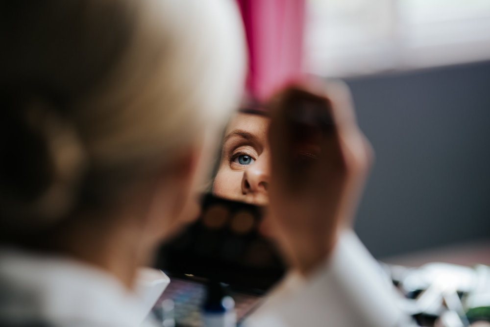  Bride's eye reflected in a small mirror as she focusedly applies makeup to her eyebrows 