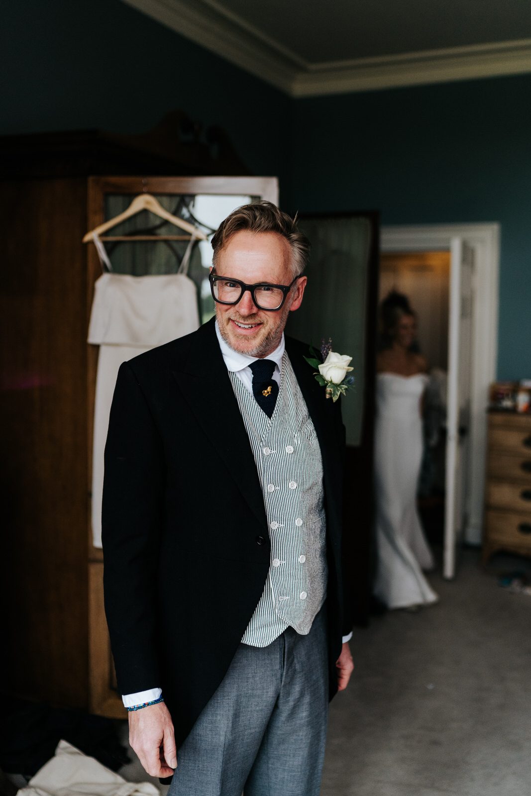  Father of the bride looks excitedly at the camera while the bride, fully dressed in her gown, enters at the back 