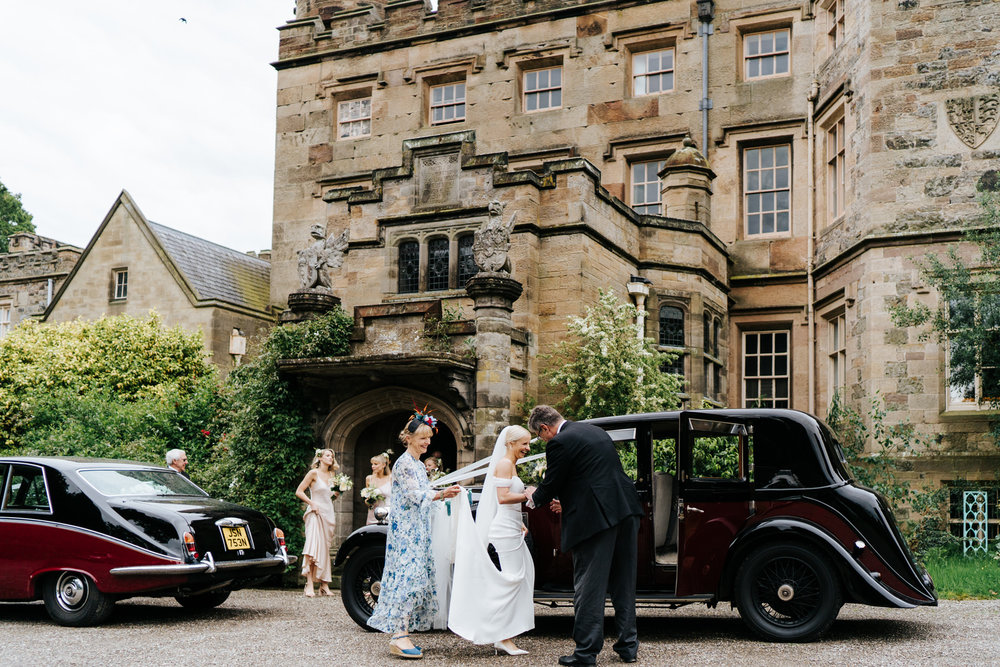  Bride, mother of the bride and bridesmaids stand against majestic Hawarden Castle venue as bride enters her wedding car 