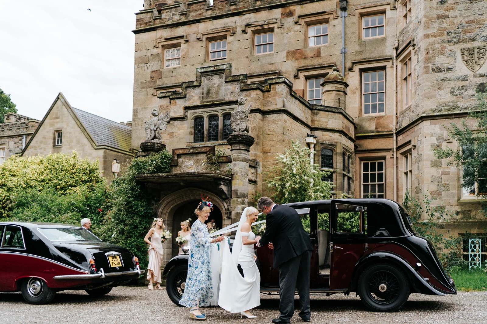 Bride, mother of the bride and bridesmaids stand against majestic Hawarden Castle venue as bride enters her wedding car