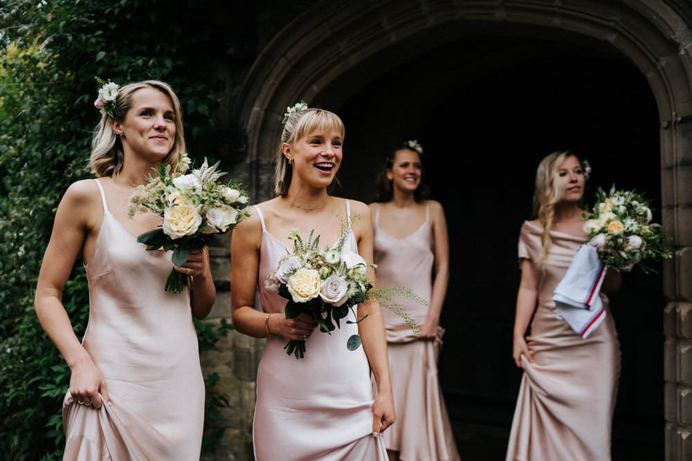  Bridesmaids hold bouquets and smile at bride, off-frame, as she enters her wedding car 