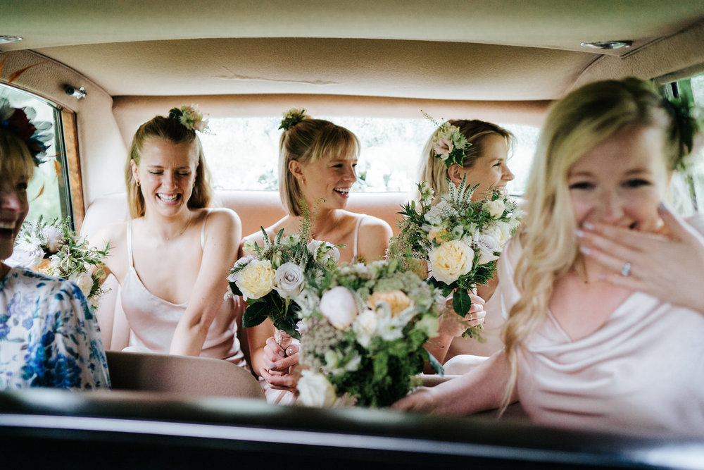  Bridesmaids and mother of the bride sit in the back of the car and smile as they are driven to wedding ceremony 