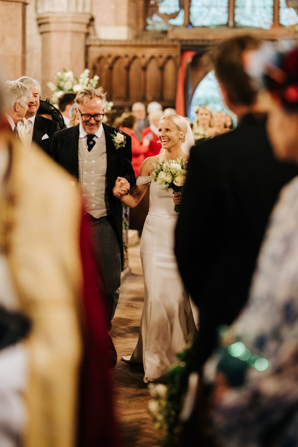  Father of the bride and bride walk down the aisle, holding hands and smiling vibrantly at Hawarden Castle Wedding 