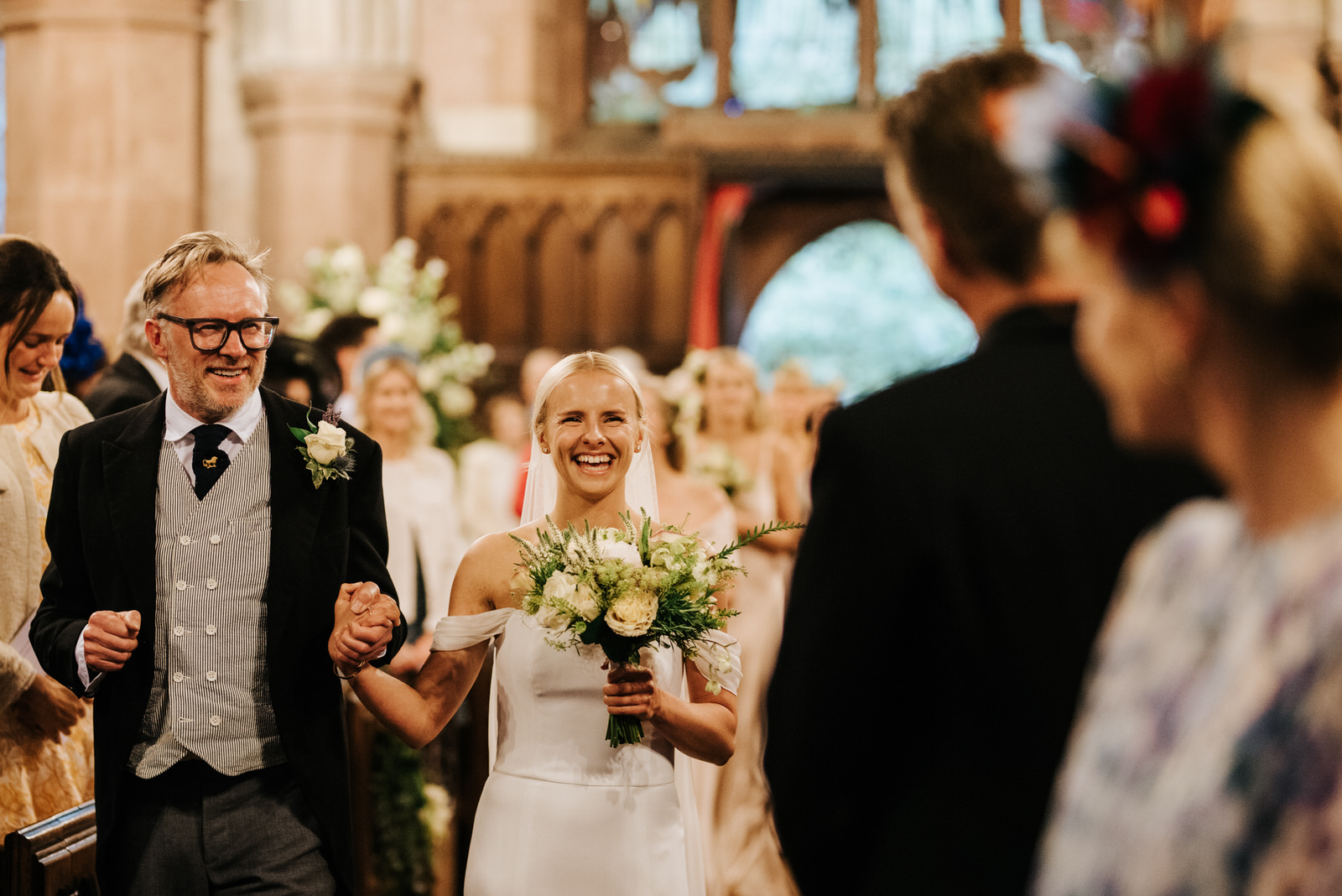  Father of the bride and bride walk down the aisle, holding hands and smiling vibrantly at Hawarden Castle Wedding 