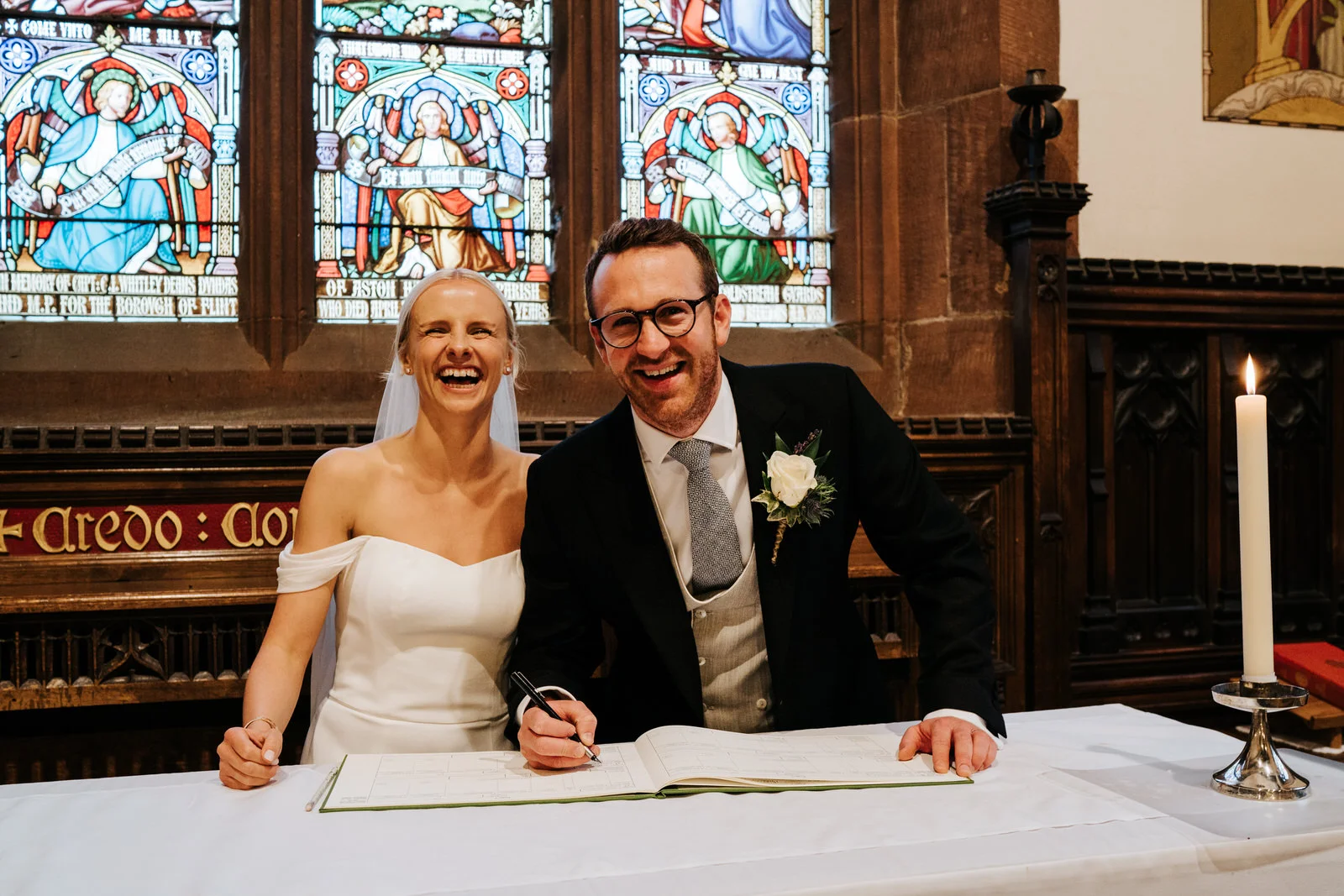 Bride and groom smile at camera while taking staged photograph of registry signing