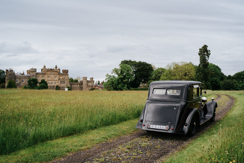  Bride and groom drive towards Hawarden Castle in beautiful, black vintage car 