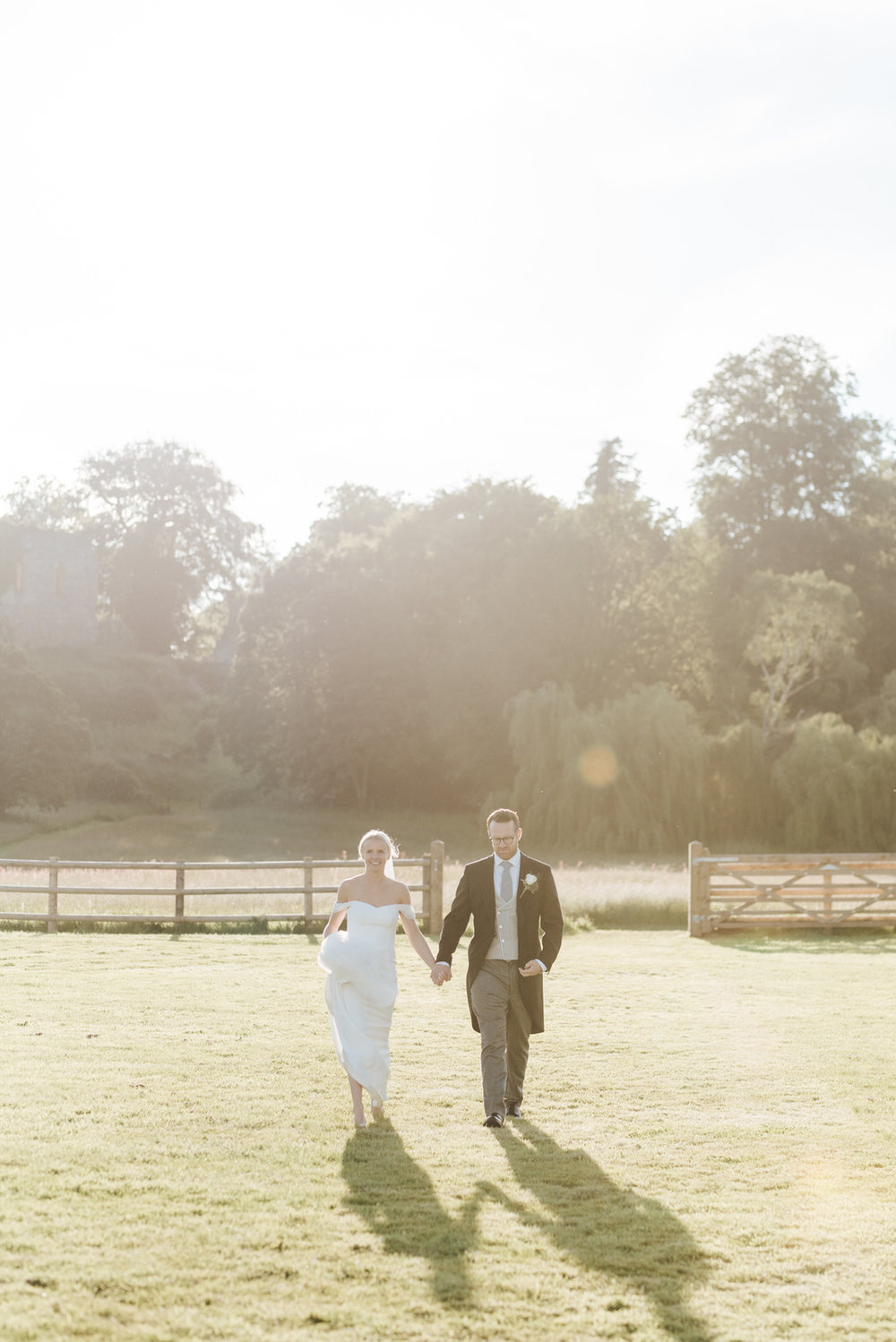  Bride and groom walk towards marquee as sun sets behind them and creates dramatic shadows 