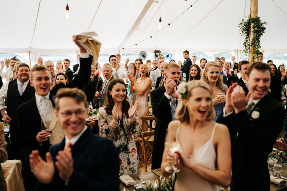  Guests cheer, smile and clap as bride and groom make their way to their table inside wedding marquee 