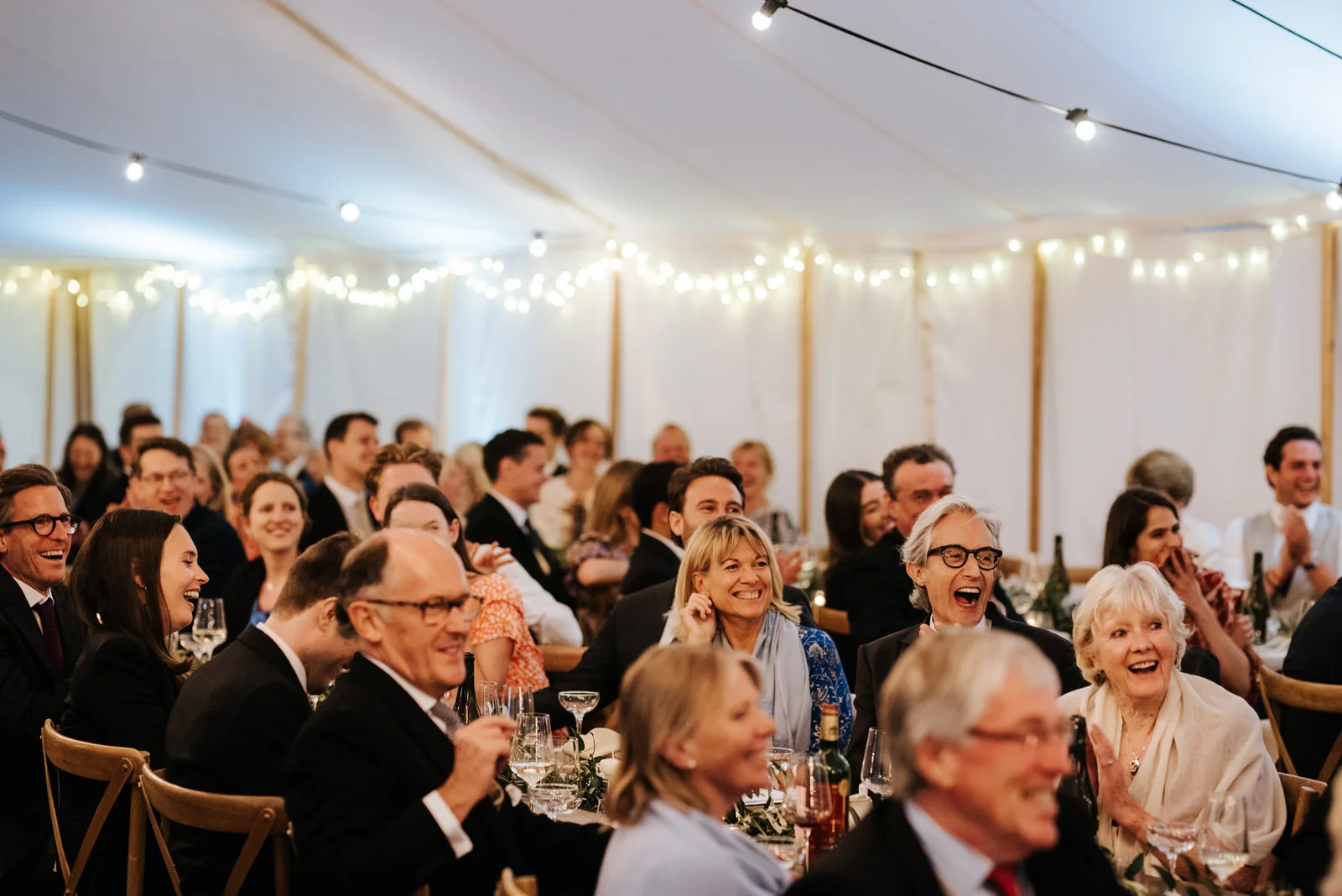 Guests clap and laugh hysterically as father of the bride, off-frame, is delivering his speech