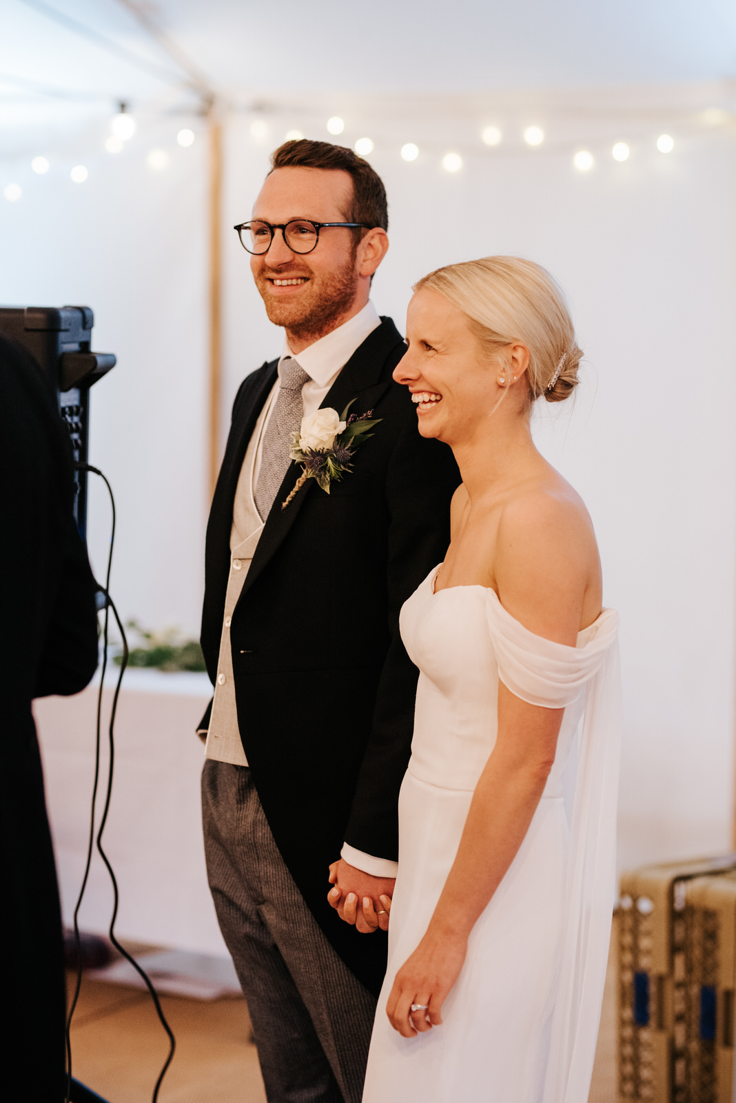 Bride and groom laugh and smile, holding hands, while fathe of the bride delivers wedding speech