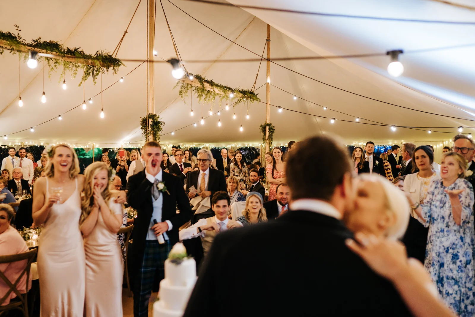 Wide photograph of bride and groom kissing and guests cheering after cutting the wedding cake