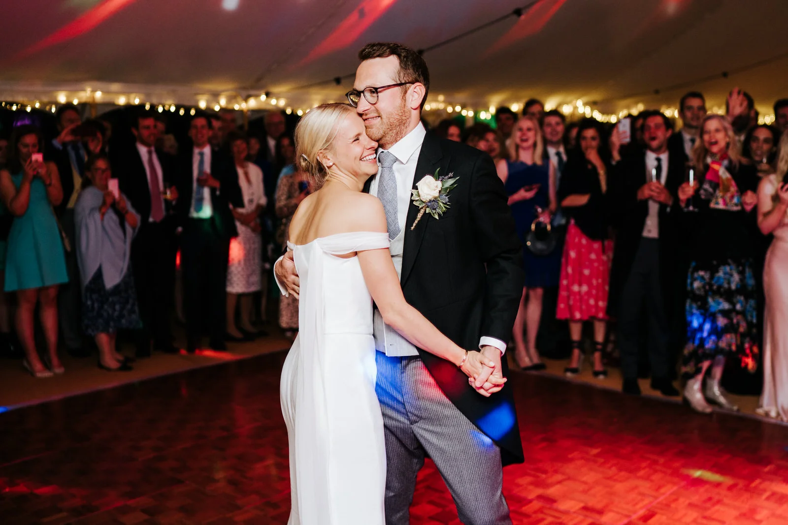 Bride and groom embrace as they have their first dance with guests cheering from the back