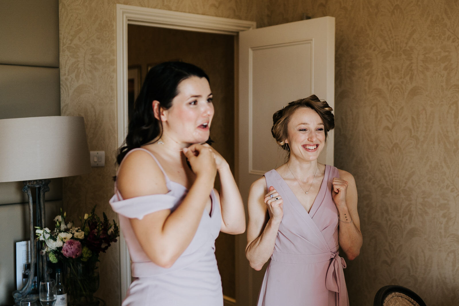  Two bridesmaids raise their hands in excitement as bridesmaid finishes getting ready 