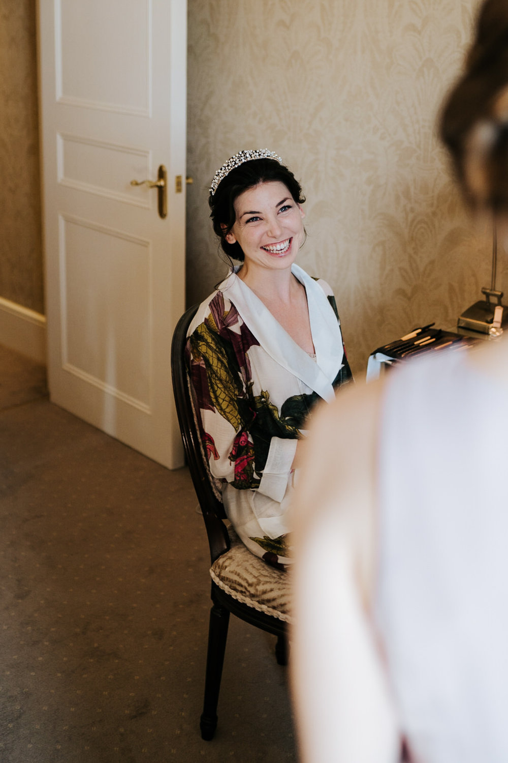  Bride sits in chair as makeup is being finished and smiles at bridesmaid 