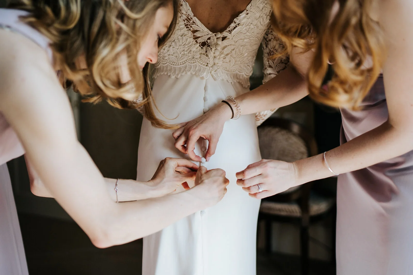 Bridesmaids finish helping bride into her dress by doing buttons up at the back