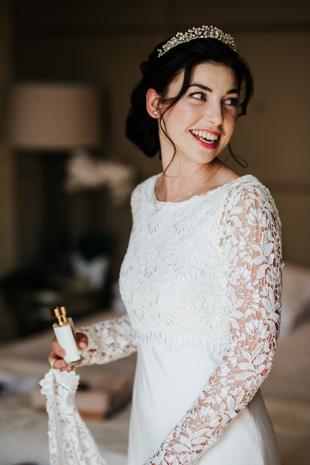  Bride turns and looks at herself in the mirror as she holds a small bottle of perfume 