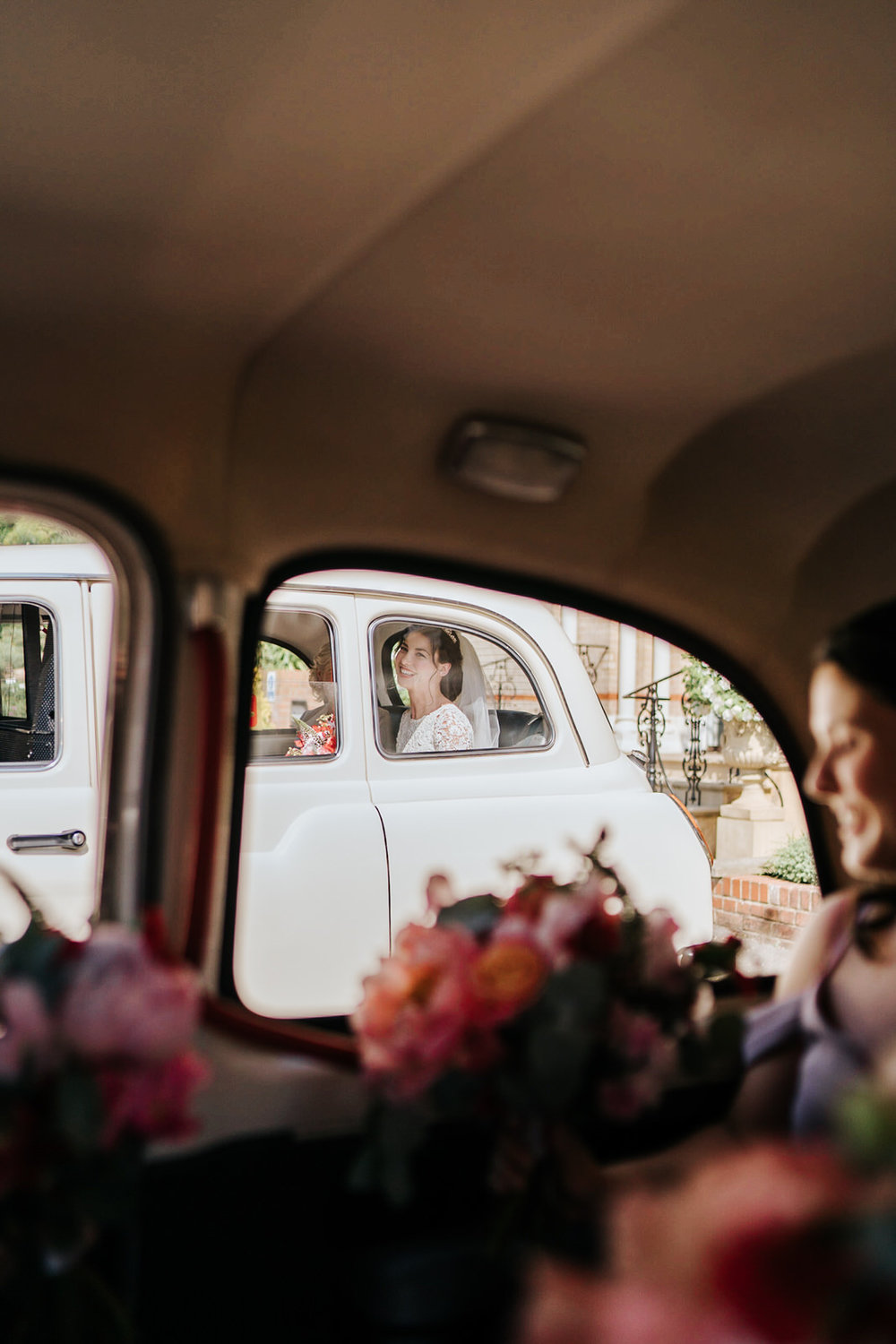  Bride looks out of the window of her wedding car and smiles at bridesmaids sitting in another car 