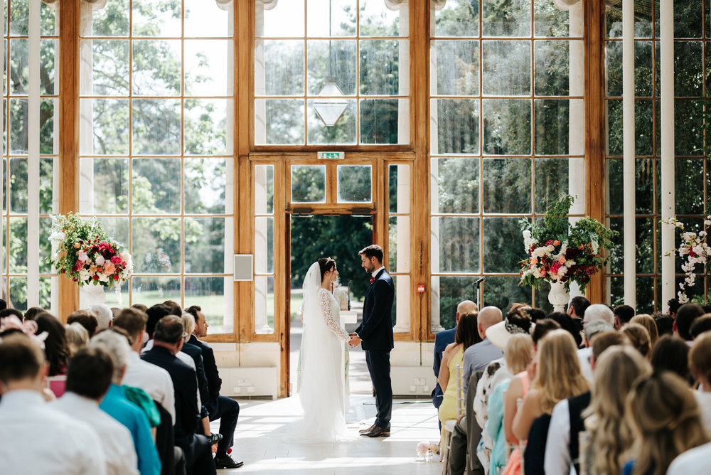  Wide photograph of bride and groom holding hands at the front of the aisle of Nash Conservatory at Kew Gardens 