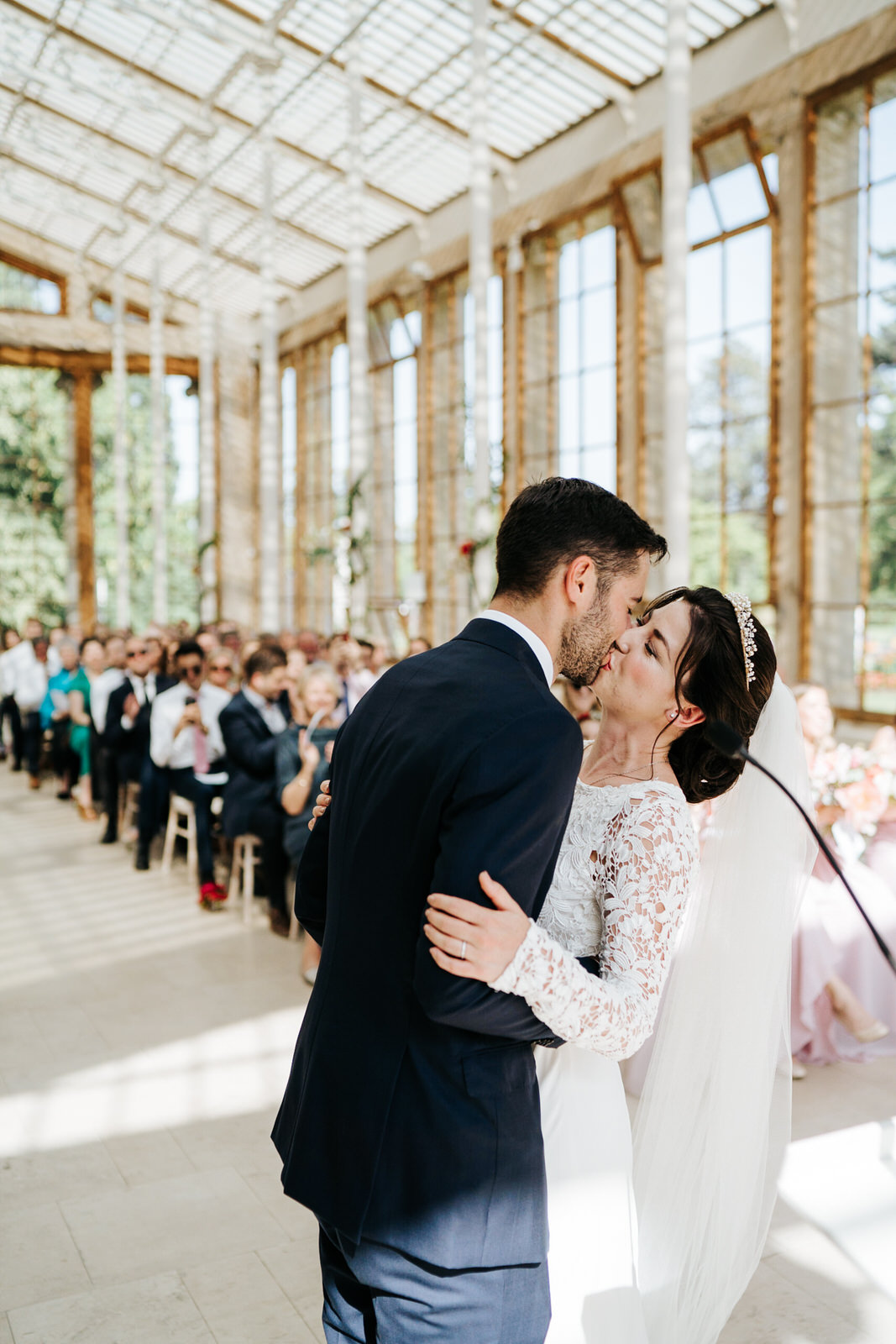  First kiss at nash conservatory wedding at Kew Gardens while guests, out of focus, look from the back. 