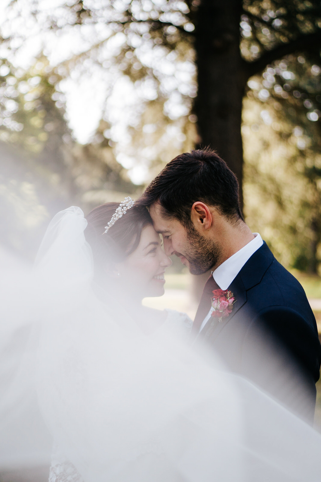  Bride and groom stand in beautiful light in Woodlands area in Kew Gardens and pose for a photograph as bride's veil takes up bottom half of the image 