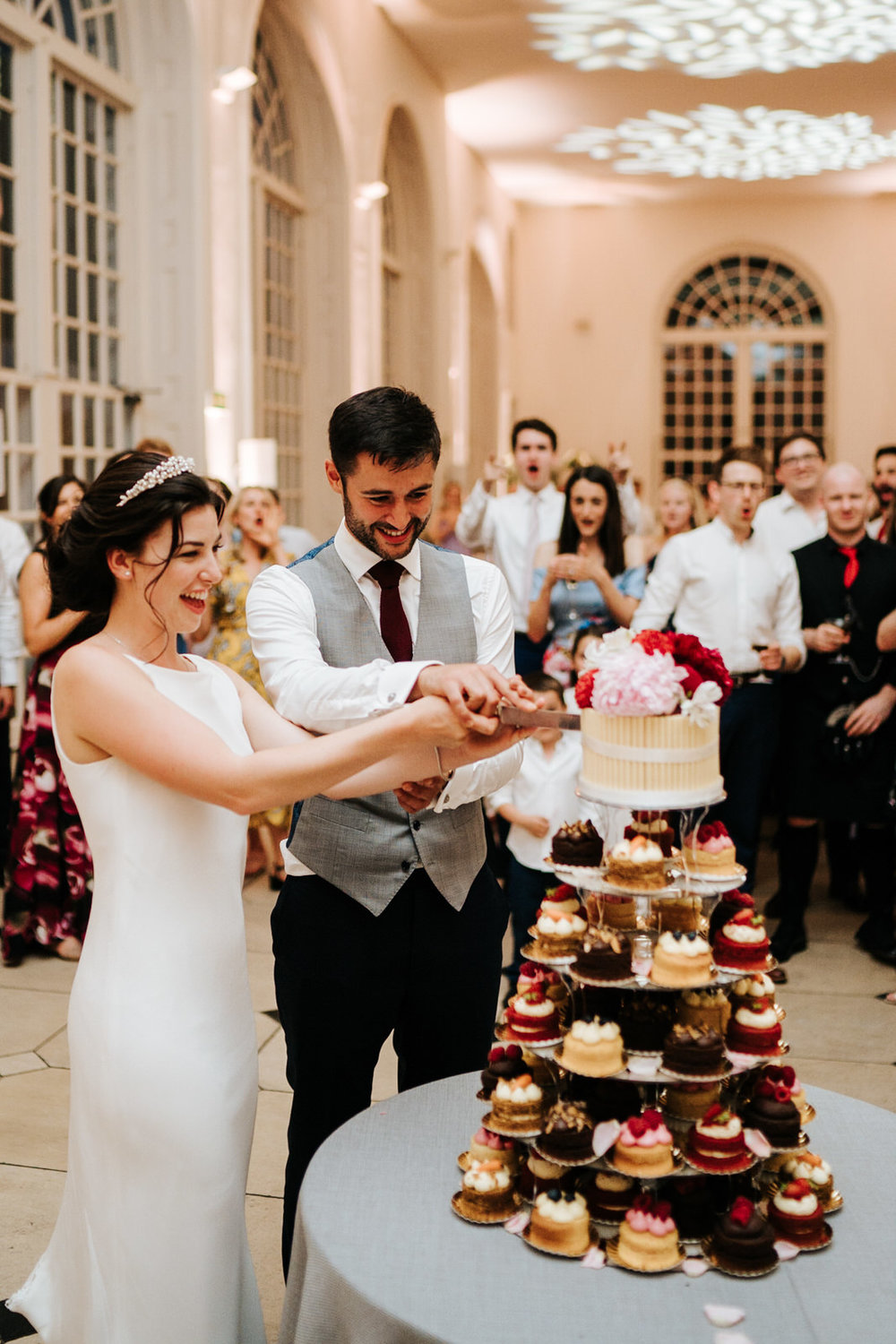  Bride and groom cut the cake as wedding guests look and smile from behind 