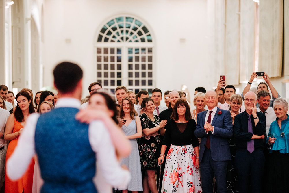  Bride and groom are out of focus on the front of the frame as guests look at them having their first dance 