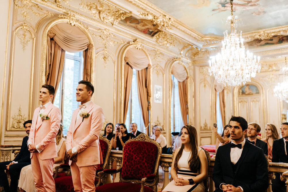  Wide photograph of both grooms standing and guests sitting behind them in ornate and golden ceremony room in Paris 