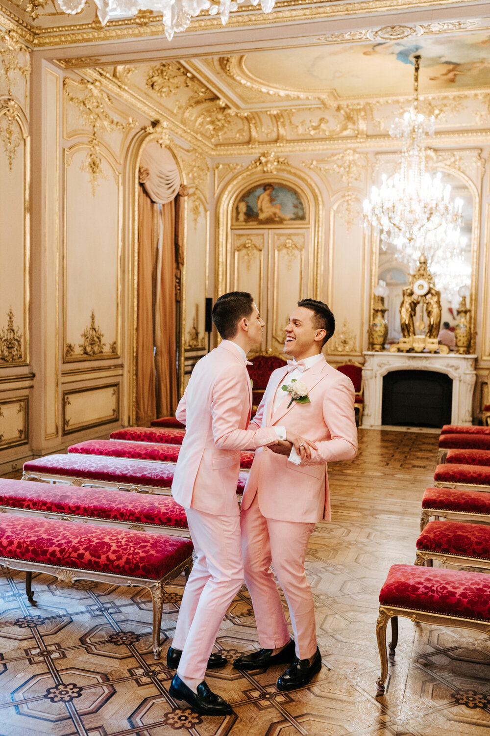  Emotional photograph of both grooms holding hands and smiling at each other in beautiful, golden and grand wedding room in Paris 