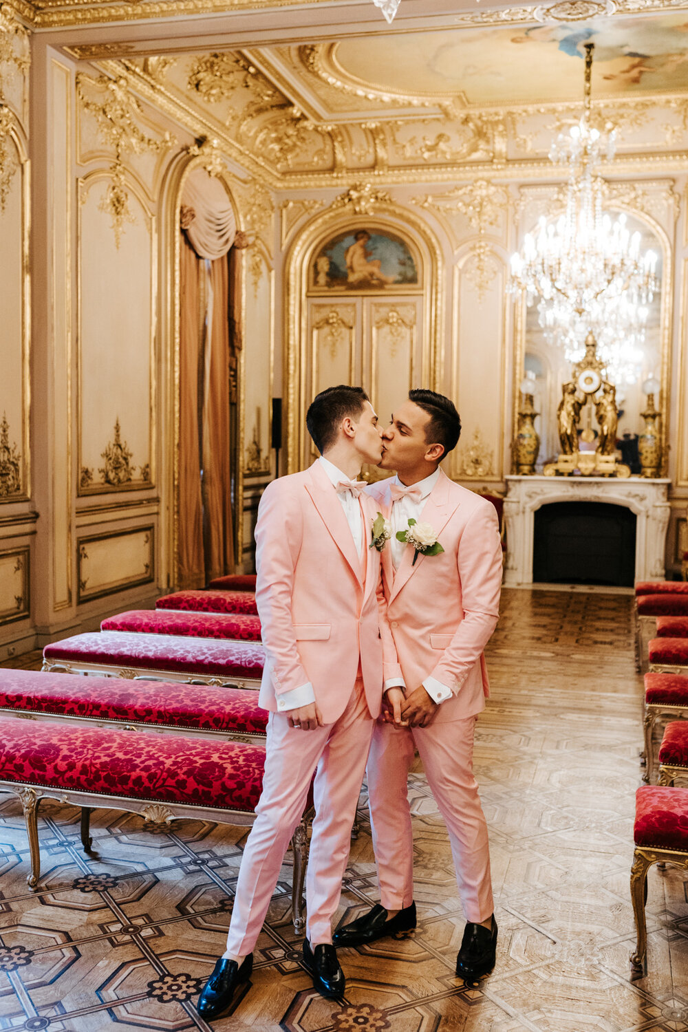  Emotional photograph of both grooms holding hands and kissing each other in beautiful, golden and grand wedding room in Paris 