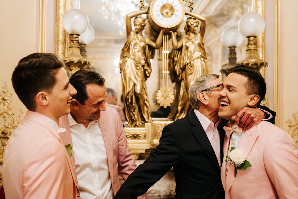  Both grooms, dad of the groom and grandfather of the groom hug and smile at each other during family photograph 