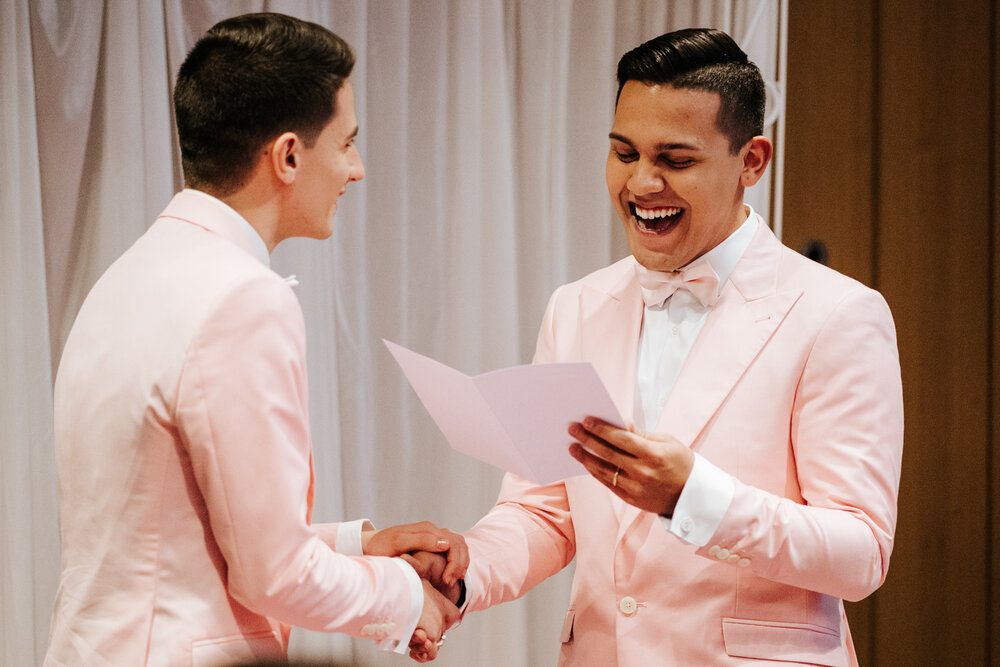  Both grooms holding hands and smiling as one of them reads his personalised wedding vows 