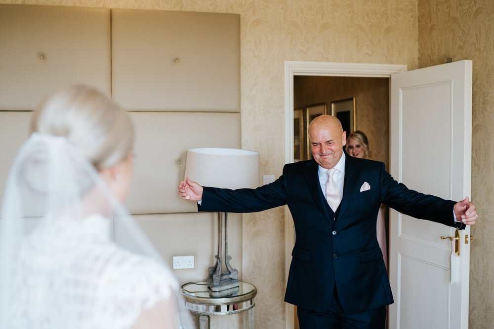 Father of the bride stretches his arms out to hug her as he sees her in her wedding dress for the first time