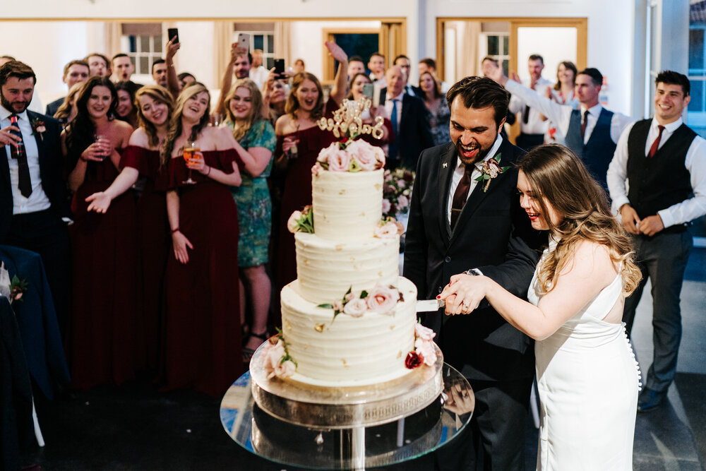 Bride and groom attempt to cut cake as guests and bridesmaids smile joyously from the background