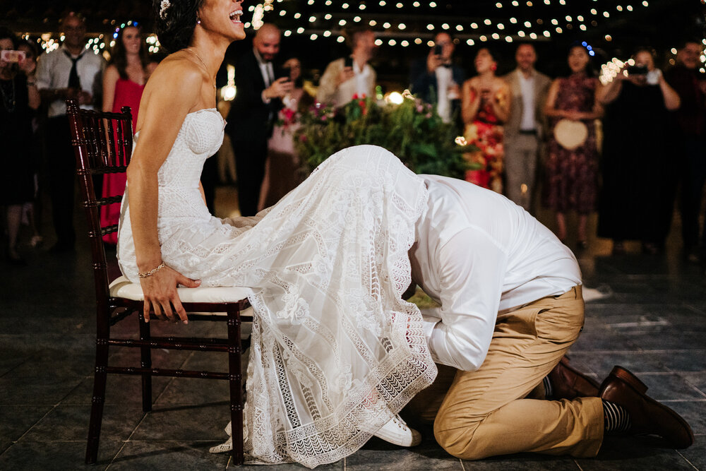 Groom's head is under bride's dress as he works on removing her garter 