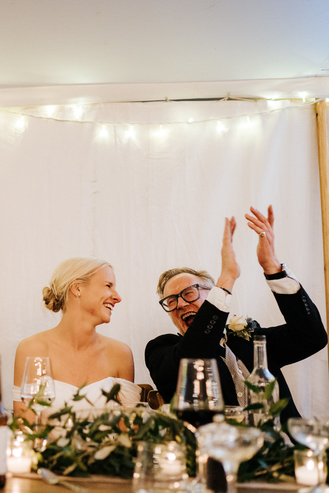  Father of the bride throws his hands into the air and claps while looking at bride, sitting right next to him and smiling 