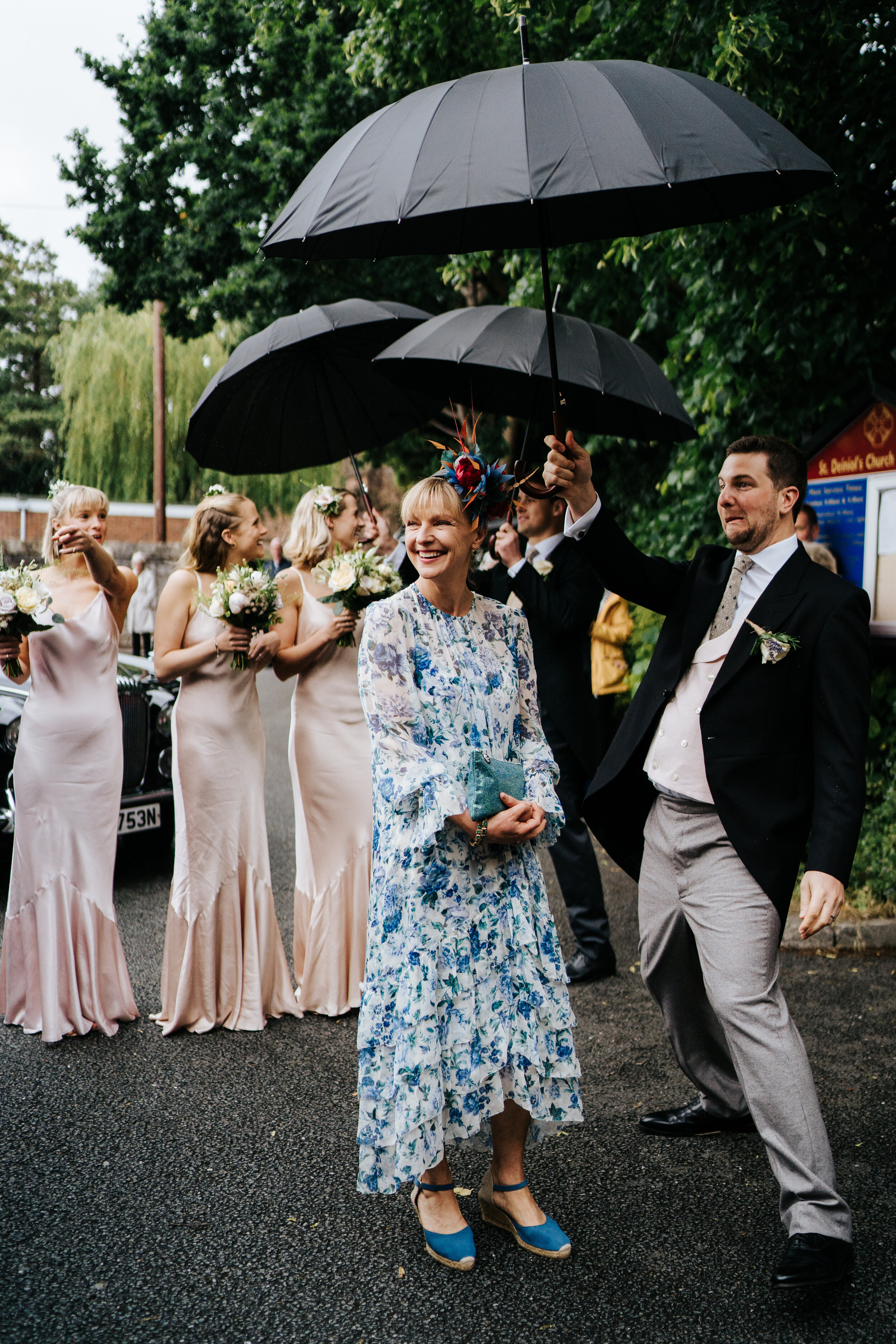 Mother of the bride and bridesmaids stand, smiling, underneath umbrellas as they wait for the bride to exit her car