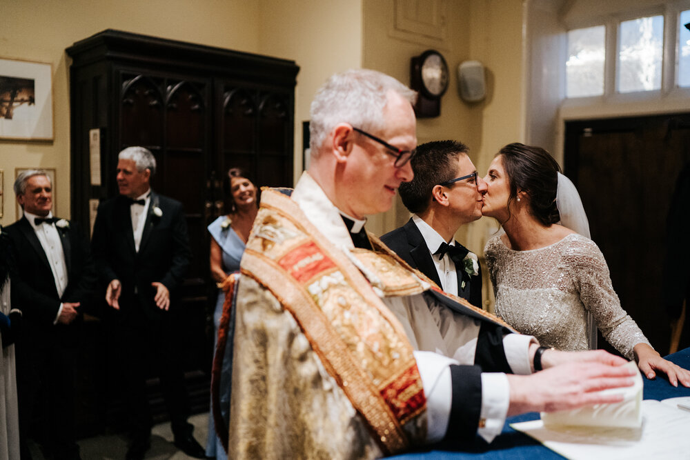 Bride and groom kiss in the background as vicar signs the register during wedding ceremony at St Pauls Knightsbridge