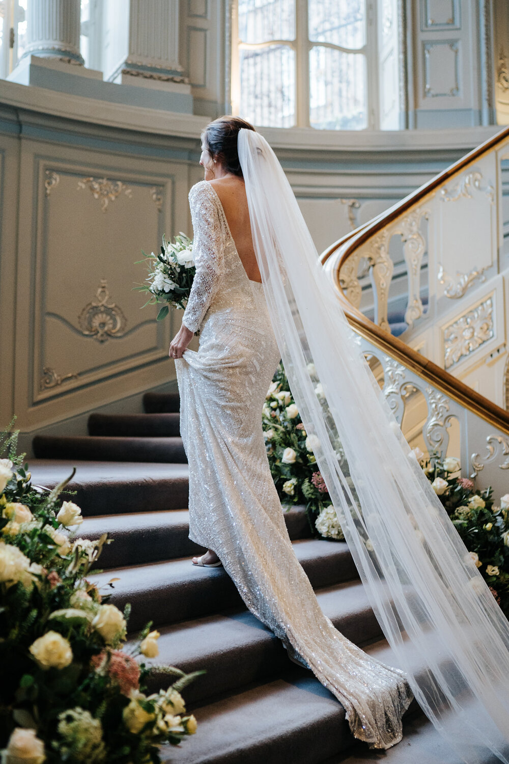 Bride walks up the decorated stairs at Savile Club London as her dress trails behind her