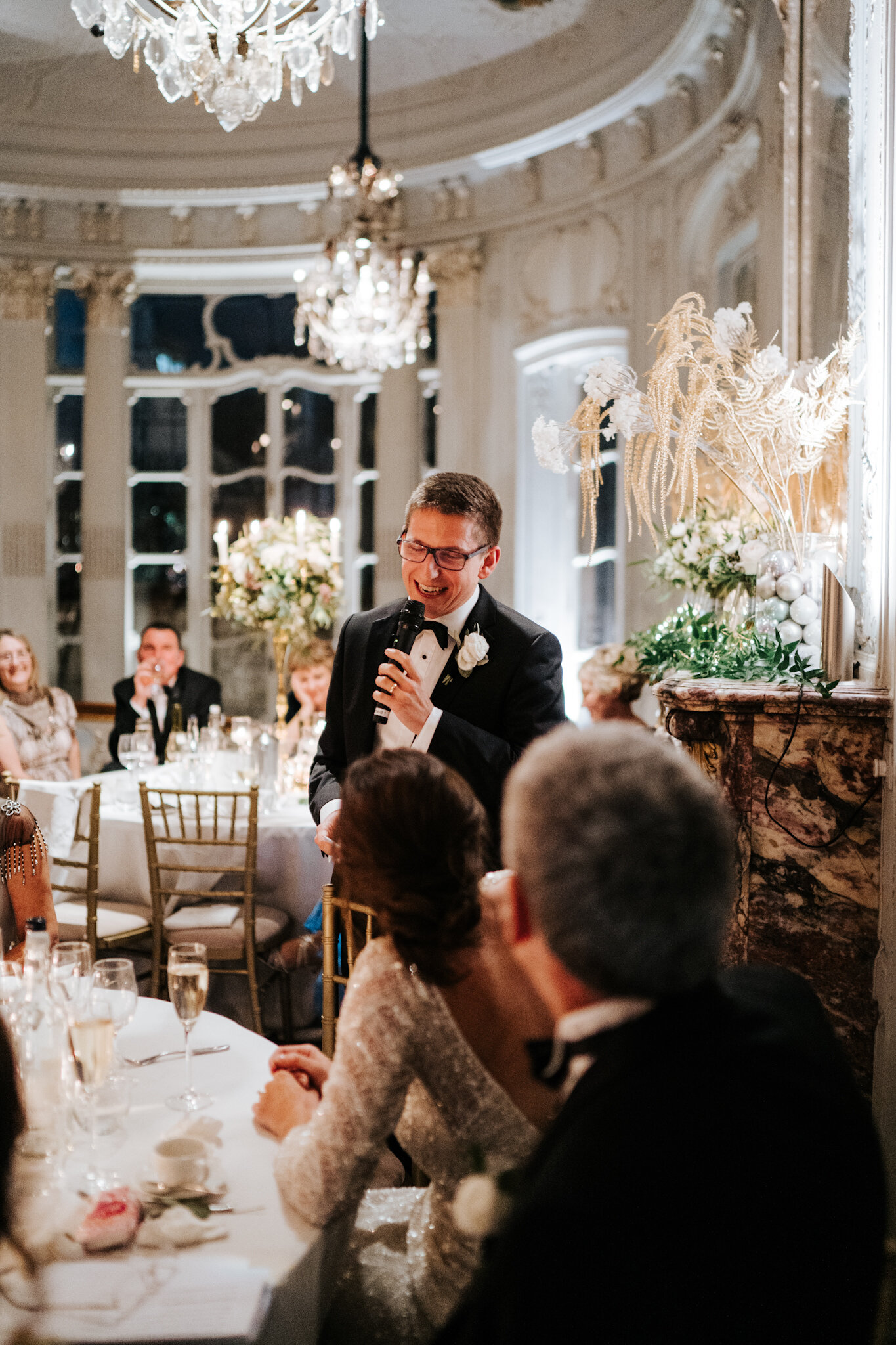 Bride covers her face with embarrassment in wide photograph of head table as father of the bride delivers speech at Savile Club London 