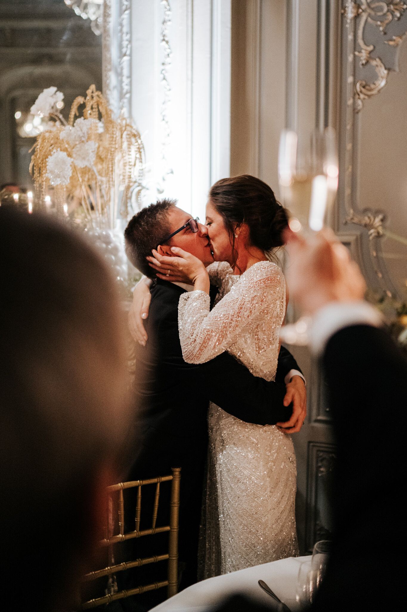 Bride and groom kiss in the background as a glass is raised in the foreground after groom concludes his speech at Savile Club London wedding