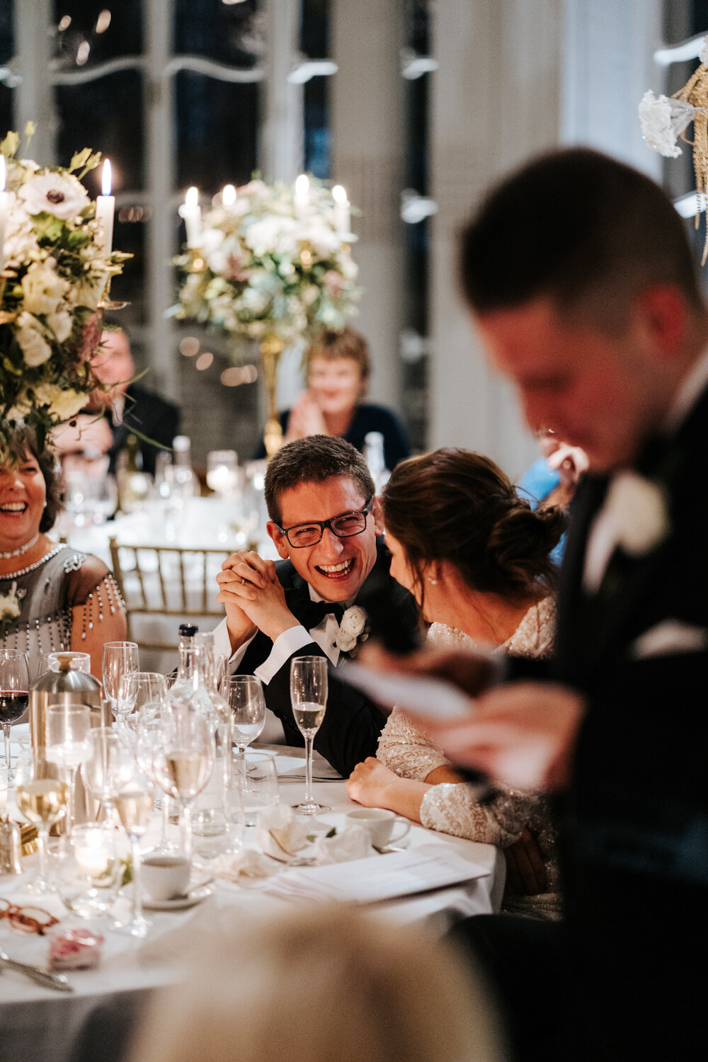 Bride and groom look at each other and smile as best man starts giving wedding speech 